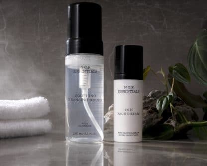Vegan & fragrance free skincare from N.C.P. Clean & Care kit with Soothing Cleansing Mousse a transparent bottle with a black cap and 24 H Face Cream a white bottle with black text and black cap.White towel and green leafs